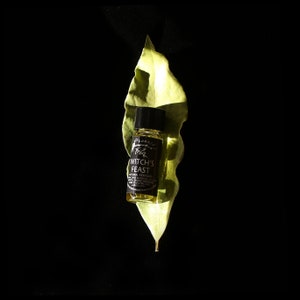 May include: A small glass bottle with a black label that reads "Witch's Feast" natural perfume. The bottle is sitting on a green leaf against a black background.