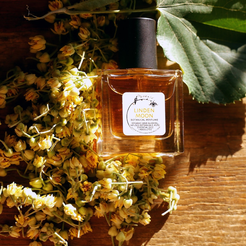 Peut inclure: Un petit flacon de parfum botanique Linden Moon en verre, avec une &eacute;tiquette brune sur laquelle est inscrit "Linden Moon Botanical Perfume Organic Cane Alcohol Infused Botanicals Pure Essential Oils Handcrafted", pos&eacute; sur une surface en bois avec des fleurs jaunes &eacute;parpill&eacute;es autour.