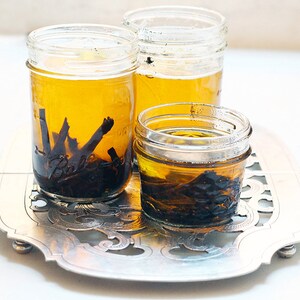 May include: Three glass jars filled with a light yellow liquid and dark brown pieces of vanilla beans. The jars are sitting on a silver tray with an ornate design.