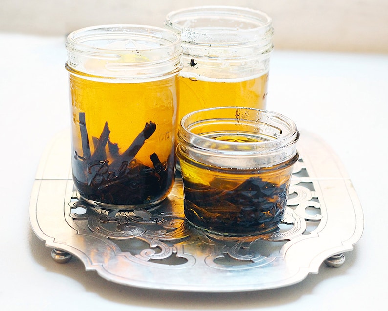 May include: Three glass jars filled with a light yellow liquid and dark brown pieces of vanilla beans. The jars are sitting on a silver tray with an ornate design.