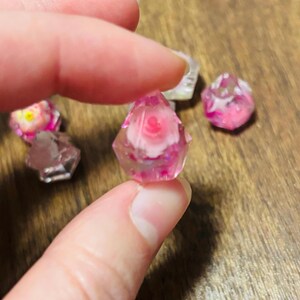 May include: A small, multifaceted, translucent pink resin bead containing a miniature white and pink flower embedded inside. The bead has a geometric, diamond-like shape with sharp edges and a glossy finish, reflecting light across its surface.