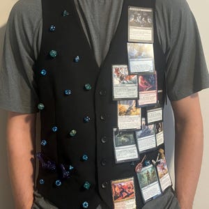 May include: A black vest adorned with numerous blue and green polyhedral dice attached to the left side and a collection of rectangular trading cards featuring various fantasy illustrations and printed text attached to the right side.