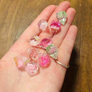 May include: A collection of small, faceted, clear resin gems containing embedded dried flowers in shades of pink, white, and green. Each piece has a geometric, diamond-like shape with sharp edges, showcasing delicate floral inclusions suspended within the transparent material.