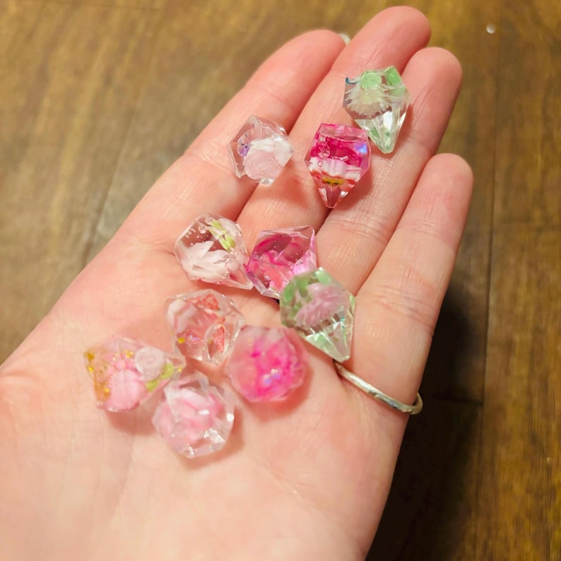 May include: A collection of small, faceted, clear resin gems containing embedded dried flowers in shades of pink, white, and green. Each piece has a geometric, diamond-like shape with sharp edges, showcasing delicate floral inclusions suspended within the transparent material.