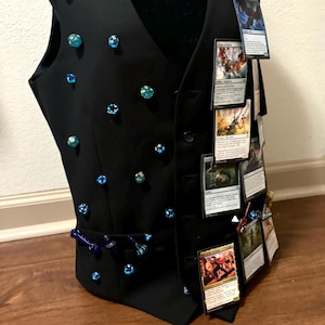 May include: A black vest adorned with numerous blue and green polyhedral dice attached to the front panel. The right side features a vertical row of multiple trading cards secured to the fabric, showcasing various illustrations and printed text.