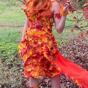 May include: A sleeveless dress covered in layered artificial autumn leaves in shades of red, orange, and yellow. The bodice features small decorative acorns and berries. A long, sheer orange fabric train extends from the side of the garment.