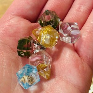 May include: A collection of six small, multi-faceted resin dice. Each die features unique inclusions, including gold leaf flakes, green moss-like textures, small white seashells, and pink accents suspended within clear, transparent geometric shapes.