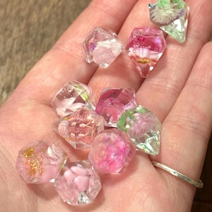 May include: A collection of small, faceted, clear resin gems containing embedded dried flowers and gold glitter. The geometric shapes feature various pink, white, and green floral inclusions suspended within the transparent material, creating a sparkling, multi-dimensional appearance across the assortment of decorative pieces.