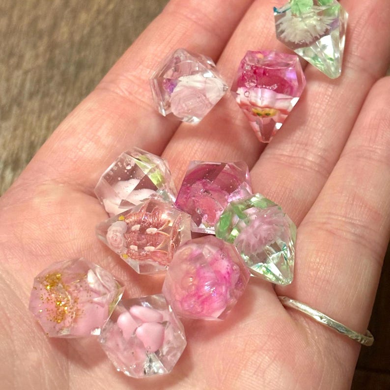 May include: A collection of small, faceted, clear resin gems containing embedded dried flowers and gold glitter. The geometric shapes feature various pink, white, and green floral inclusions suspended within the transparent material, creating a sparkling, multi-dimensional appearance across the assortment of decorative pieces.