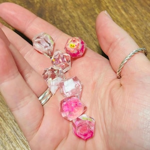 May include: A collection of six small, multi-faceted resin dice featuring embedded floral elements in shades of pink, white, and green. Each die has a clear, glossy finish with distinct geometric shapes, showcasing delicate botanical inclusions suspended within the transparent material.