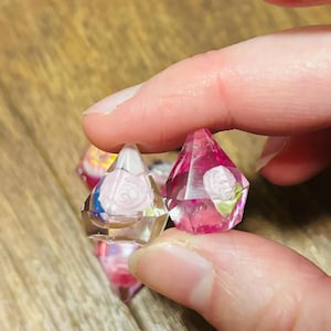 May include: Two faceted, diamond-shaped resin charms featuring embedded miniature white rose floral details. One charm is clear with a pale pink rose, while the other is translucent pink with a white rose. Both have sharp, geometric edges.