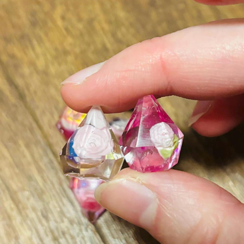 May include: Two faceted, diamond-shaped resin charms featuring embedded miniature white rose floral details. One charm is clear with a pale pink rose, while the other is translucent pink with a white rose. Both have sharp, geometric edges.