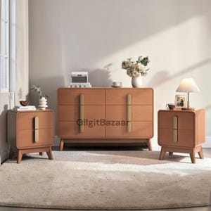 May include: A bedroom furniture set featuring a dresser and two nightstands. The wooden furniture has a light brown finish with gold-colored handles. The nightstands have a lamp and a framed photo. A vase of flowers sits atop the dresser.
