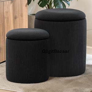 May include: Two black, cylindrical storage ottomans with ribbed fabric texture. One ottoman is larger than the other, both have a flat, circular lid. The ottomans are on a neutral-colored rug.