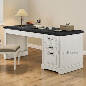 May include: A two-toned wooden desk with a black top and white base. The desk features three drawers and a cabinet door on the right side. A lamp, books, and a small box are on the desk. A chair is in the foreground.