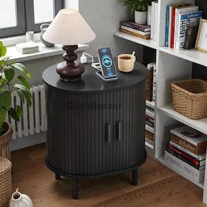 May include: A dark gray, round bedside table with a ribbed design and two doors. A brown lamp with a beige shade sits on top, along with a phone charging on a wireless charger. A small cup of coffee is also on the table. The table is in a room with a window and a bookshelf.