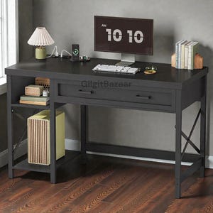 May include: A dark gray desk with a drawer, a shelf, and metal legs. A computer monitor displays "10 10" and a keyboard sits on the desk. A lamp, books, and a charging station are also on the desk. The desk is on a wooden floor.