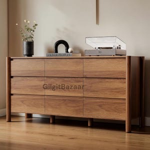 May include: A wooden dresser with nine drawers, featuring a warm brown finish. The dresser has a rectangular shape with rounded edges and sits on short legs. A record player and decorative items are on top. The text "GilgitBazaar" is visible.