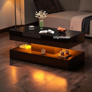 May include: A modern black coffee table with a glossy finish, featuring a lower shelf with warm lighting. The table is rectangular and has decorative items on top, including a vase, book, and fruit bowl. The text "GilgitBazaar" is visible on the table.