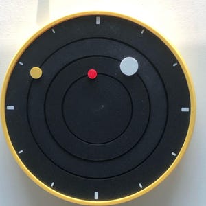 May include: A contemporary wall clock with a black face and a yellow surround. The clock has a circular design with concentric rings and small coloured dots representing planets. White rectangular markers indicate the hours.