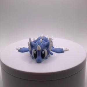 Könnte beinhalten: Hellblaue Drachenfigur mit weißen Akzenten, liegend auf einer weißen Oberfläche. Der Drache hat große schwarze Augen, ein weißes Horn und weiße Stacheln auf Rücken und Flügeln. Eine skurrile Fantasiefigur.