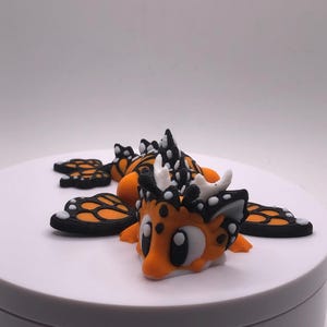 May include: An orange and black creature figurine with large eyes, white horns, and black and white accents. The figurine has four orange and black wings. The creature is lying on a white surface.