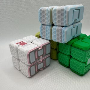 May include: Three small, colourful, cube-shaped fidget toys. The top cube is light blue with a white honeycomb pattern. The middle cube is yellow with a white honeycomb pattern. The bottom cube is green with a white honeycomb pattern.