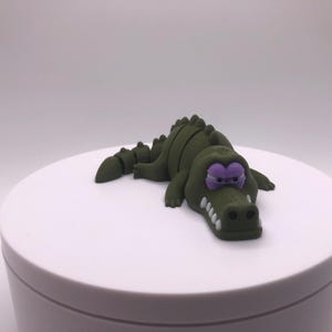 May include: A green alligator figurine with purple eyes and white teeth. The alligator is lying down on a white surface. The figurine has a cartoonish design.