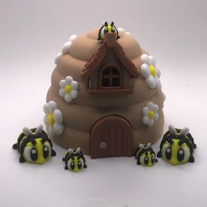 May include: A whimsical, brown beehive-shaped decoration with a small house-like structure, adorned with white and yellow flowers. Several cartoon bees in yellow and black surround the hive, creating a playful, nature-inspired scene.