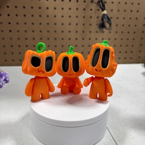 Peut inclure: Trois figurines orange en forme de citrouille avec des tiges vertes et des yeux noirs. Les figurines sont posées sur une plate-forme circulaire blanche.
