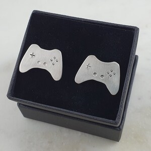 May include: Two silver-colored cufflinks in the shape of a video game controller, displayed in a black velvet box.