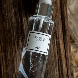 May include: A clear glass spray bottle with a silver cap, filled with a liquid. The label reads "Lemongrass Hydrosol" with the initials "AV". The bottle is set against a rustic, dark wood background.