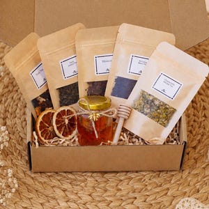 May include: A cardboard gift box containing five kraft paper pouches of loose-leaf tea, a small glass jar of amber honey with a bee charm, and a wooden honey dipper. Two dried orange slices are placed in the front of the box with paper filler.