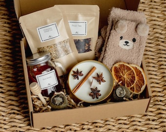 May include: A gift box filled with relaxation items. It includes a jar of honey, herbal tea, fruit tea, a candle with star anise and a cinnamon stick, dried orange slices, and a pair of bear-shaped socks. The box is brown and sits on a woven surface.