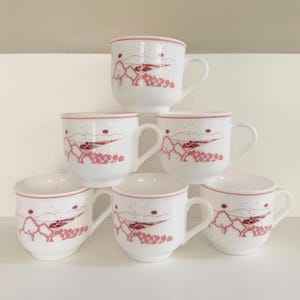 May include: A set of six white ceramic mugs stacked in a pyramid. Each mug features a red landscape design with mountains, trees, and a house. The rims and handles are also red.