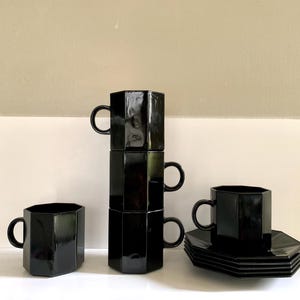 May include: A set of black, geometric-shaped coffee cups and saucers. The cups are stacked in a tower of three, with two additional cups and a stack of saucers arranged nearby. The glossy finish reflects light, highlighting the angular design.