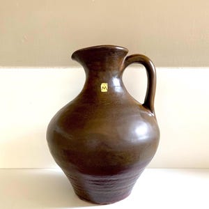 Può includere: Una brocca in ceramica marrone con manico e beccuccio. La brocca ha un corpo bulboso e una finitura liscia e lucida. Un piccolo adesivo quadrato con un logo è attaccato sul davanti. La brocca viene probabilmente utilizzata per servire bevande o come oggetto decorativo.