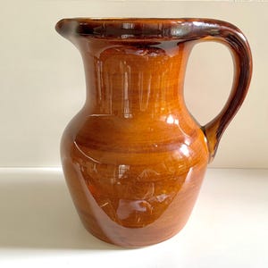 Pode incluir: Um jarro de cerâmica vidrada com corpo arredondado e alça curva. O jarro é de cor castanha quente com detalhes castanhos mais escuros e acabamento brilhante. É um item decorativo.