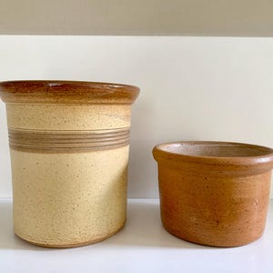 Op de afbeelding: Twee keramische keukenpotten. De grotere is beige met bruine accenten en horizontale strepen. De kleinere is effen bruin van kleur. Beide hebben een rustieke, handgemaakte uitstraling.