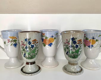 Może przedstawiać: Zestaw sześciu ceramicznych filiżanek do kawy z kwiatowymi wzorami. Filiżanki mają nóżkę i są ozdobione niebiesko-białymi kratkami i kolorowymi kwiatami.