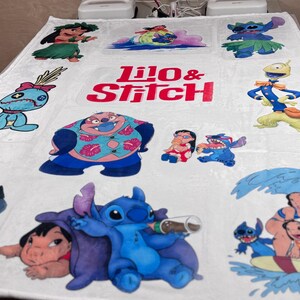 May include: White blanket featuring various colorful images of characters from the animated film "Lilo & Stitch." The blanket includes the text "Lilo & Stitch" in red and pink. The blanket is rectangular.