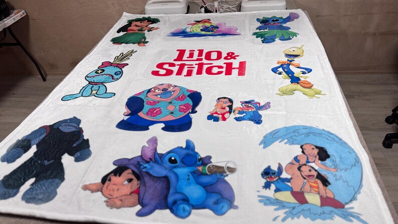 May include: White blanket featuring various colorful images of characters from the animated film "Lilo & Stitch." The blanket includes the text "Lilo & Stitch" in red and pink. The blanket is rectangular.