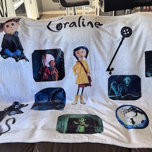 May include: White Coraline-themed blanket featuring various character illustrations and the film's title. The design includes images of Coraline, a black cat, a key, and other characters from the movie. The blanket is displayed on a dark brown leather couch.