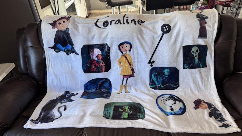 May include: White Coraline-themed blanket featuring various character illustrations and the film's title. The design includes images of Coraline, a black cat, a key, and other characters from the movie. The blanket is displayed on a dark brown leather couch.