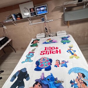 May include: White blanket featuring colorful cartoon characters from Lilo & Stitch. The blanket has the words "Lilo & Stitch" in red and white. Various characters are depicted in blue, pink, and green.