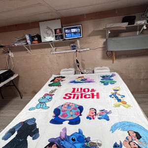 May include: White blanket featuring various colorful illustrations of characters from the animated film "Lilo & Stitch." The blanket has the words "Lilo & Stitch" in red and pink. The blanket is on a gray surface.