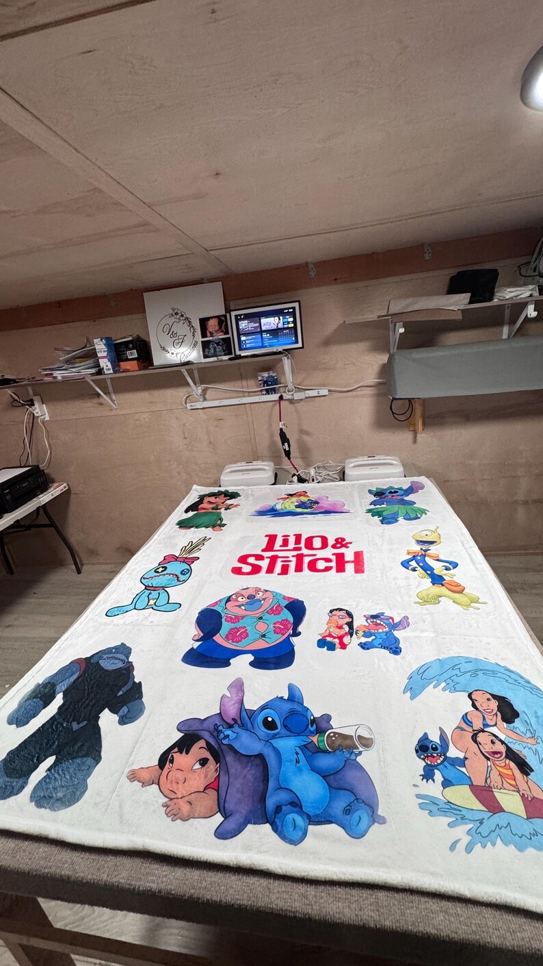 May include: White blanket featuring various colorful illustrations of characters from the animated film "Lilo & Stitch." The blanket has the words "Lilo & Stitch" in red and pink. The blanket is on a gray surface.