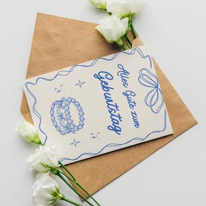 May include: A birthday card with blue illustrations and text, placed inside a brown envelope. White flowers with green stems are arranged around the card and envelope on a white surface.