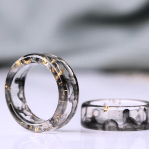 Könnte beinhalten: Zwei transparente Resinringe mit schwarzen und goldenen Flocken. Die Ringe sind dick und haben eine abgerundete Form.