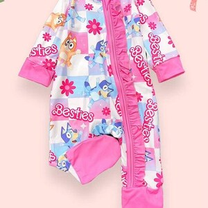 Może przedstawiać: R&oacute;żowo-białe śpioszki dla dziecka z postaciami z kresk&oacute;wek, kwiatami i napisem "Besties". Śpioszki mają zamek błyskawiczny, r&oacute;żowe wykończenia i dołączone stopy. Wz&oacute;r zawiera wz&oacute;r w szachownicę.